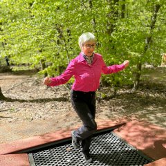 Eine Person balanciert auf einer federnden Outdoor-Station im Wald. Sie steht mit ausgebreiteten Armen auf einer schwarzen, elastischen Fläche, die von roten Platten umgeben ist. Die Trainingsstation befindet sich auf einem Waldplatz mit Laubbäumen und grünem Bewuchs im Hintergrund.
