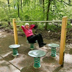 Eine Person trainiert an einer Outdoor-Fitnessstation im Wald. Sie sitzt auf einer runden, federnden Sitzfläche und hält die Hände hinter dem Kopf, während sie eine Übung für die Bauchmuskulatur ausführt. Die Station besteht aus Holzpfosten, Metallstangen und mehreren runden Federplattformen und steht auf einem befestigten Waldplatz mit dichtem grünem Baumbestand im Hintergrund.
