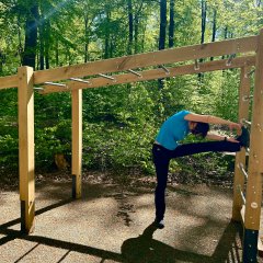 Eine Person führt eine Dehnübung an einer hölzernen Outdoor-Trainingsstation im Wald aus. Sie steht auf einem Bein und streckt das andere Bein seitlich auf eine Metallstange, während sie sich nach vorne beugt. Die Station besteht aus Holzpfosten und Metallgriffen und steht auf einem befestigten Waldplatz mit dichtem grünem Baumbestand im Hintergrund.