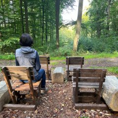 herz-Kreislauf-Pfad Lahnstein Eine Frau sitzt mit dem Rücken zur Kamera auf einem Holzstuhl an einem Ruheplatz mitten im grünen Laubwald. Um sie herum stehen mehrere weitere einfache Holzstühle sowie massive Holzbalken als Sitzgelegenheiten. Die Frau blickt in den dicht bewachsenen Wald. Hinter dem Sitzplatz verläuft ein schmaler Waldweg. Sonnenlicht fällt zwischen den hohen Bäumen hindurch und beleuchtet Teile des Waldes.