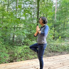 Herz-Kreislauf-Pfad Lahnstein Eine Frau steht auf einer großen Holzplattform mitten in einem grünen Laubwald und führt eine Yogaübung aus. Sie steht auf einem Bein, während der Fuß des angewinkelten anderen Beins an der Innenseite des Standbeins ruht. Ihre Handflächen hält sie vor der Brust aneinander. Die Frau blickt konzentriert nach vorne. Die von hohen Bäumen und dichtem Grün umgebene Plattform dient als ruhiger Ort für Bewegung und Entspannung in der Natur.
