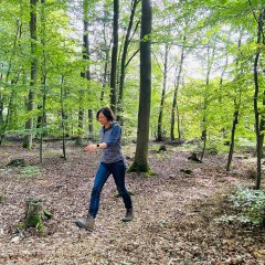 Herz-Kreislauf-Pfad Lahnstein Eine Frau bewegt sich durch einen grünen Laubwald und führt dabei eine Bewegungs- oder Achtsamkeitsübung aus. Sie macht einen großen Schritt nach vorne und hält die Arme locker vor dem Körper. Der Waldboden ist mit trockenem Laub, Ästen und einzelnen Baumstümpfen bedeckt. Hohe Bäume und dichtes grünes Blattwerk umgeben die Frau, während Sonnenlicht durch die Baumkronen fällt.