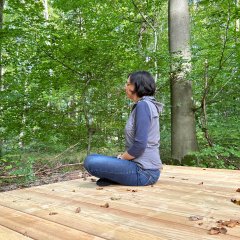 Heiz-Kreislauf-Pfad Lahnstein Eine Frau sitzt mit gekreuzten Beinen auf einer großen Holzplattform mitten in einem dicht bewachsenen, grünen Laubwald. Sie ist im Seitenprofil zu sehen, hält die Hände entspannt im Schoß und blickt ruhig in den Wald. Die Szene vermittelt eine ruhige, naturnahe Atmosphäre.