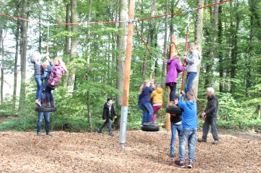 Kinderheilwald Lahnstein Mehrere Kinder spielen auf einem Bewegungs- und Kletterparcours mitten im grünen Laubwald. Sie stehen in kleinen Gruppen auf frei schwingenden Autoreifen und halten sich an roten Seilen fest, die zwischen hohen Holzpfosten gespannt sind. Einige Kinder unterstützen sich gegenseitig beim Balancieren, während zwei Erwachsene die Gruppe begleiten und Hilfestellung geben. Der Waldboden unter dem Parcours ist mit Holzspänen bedeckt.