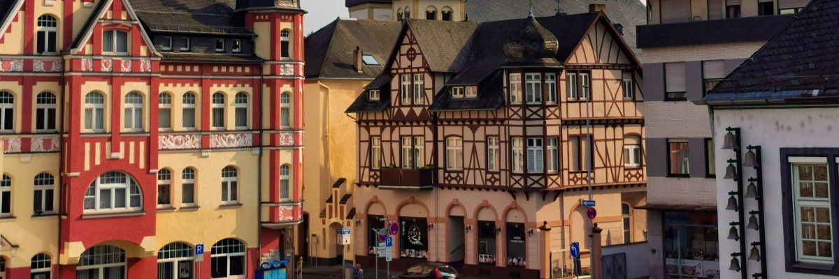 Mehrere historische Gebäude in Lahnstein, darunter ein auffälliges Fachwerkhaus mit vielen Fenstern und ein bunt verziertes Gebäude mit roten und gelben Fassadenelementen. Im Hintergrund ragt ein Kirchturm mit spitzem Dach in den Himmel. Die Szene ist bei Tageslicht aufgenommen, mit leicht bewölktem Himmel, und im Vordergrund sind Straßen und vereinzelte Verkehrsschilder zu sehen.