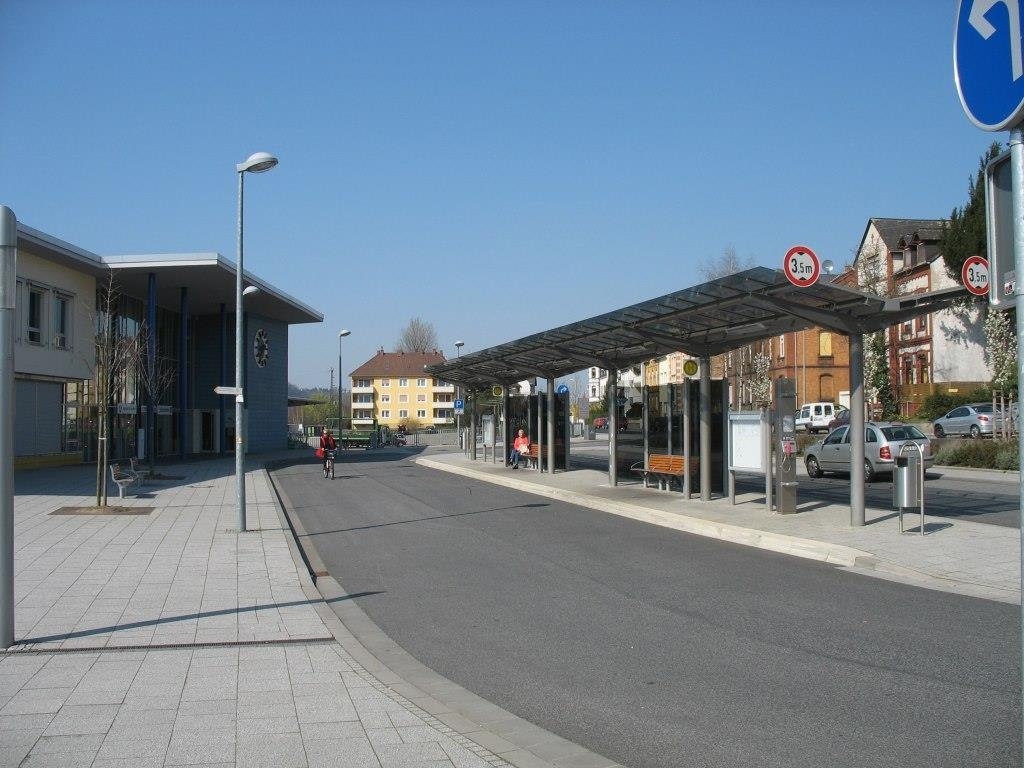 Bahnhofsvorplatz in Niederlahnstein mit Bushaltestelle an einer breiten, sauberen Straße an einem sonnigen Tag. Rechts steht ein modernes, überdachtes Wartehäuschen mit Glaswänden und Sitzbänken, unter dem einige Personen sitzen. Links befindet sich ein modernes Gebäude mit großen Fenstern. Im Hintergrund sind Wohnhäuser und parkende Autos zu sehen. Eine Person fährt mit dem Fahrrad die Straße entlang. Verkehrsschilder zeigen eine Höhenbegrenzung von 3,5 Metern an.