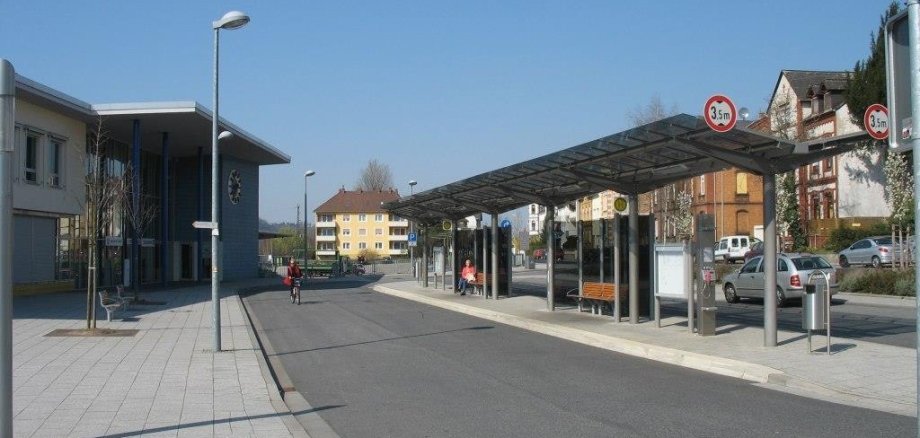 Bahnhofsvorplatz in Niederlahnstein mit Bushaltestelle an einer breiten, sauberen Straße an einem sonnigen Tag. Rechts steht ein modernes, überdachtes Wartehäuschen mit Glaswänden und Sitzbänken, unter dem einige Personen sitzen. Links befindet sich ein modernes Gebäude mit großen Fenstern. Im Hintergrund sind Wohnhäuser und parkende Autos zu sehen. Eine Person fährt mit dem Fahrrad die Straße entlang. Verkehrsschilder zeigen eine Höhenbegrenzung von 3,5 Metern an.