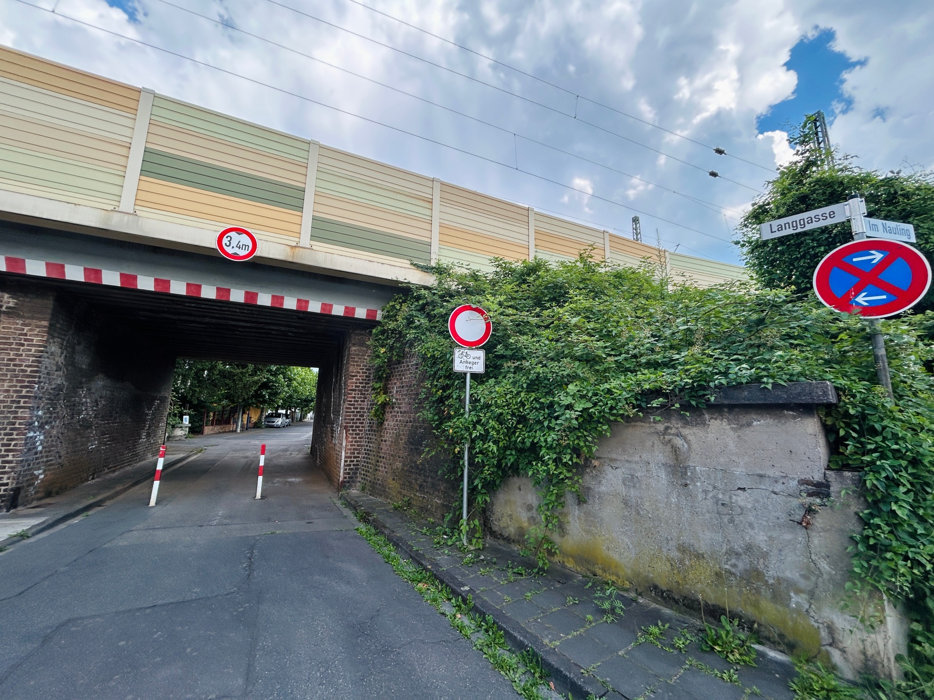 Ansicht der Bahnunterführung in einer Straße, der Langgasse in Lahnstein, mit der darüber verlaufenden Bahnstrecke. Über der Unterführung weist ein Schild auf eine maximale Durchfahrtshöhe von 3,4 Metern hin. Rechts sind die Straßenschilder „Langgasse“ und „Im Nauling“ sowie ein eingeschränktes Halteverbot zu sehen. Die Durchfahrt wird durch zwei rot-weiße Poller verengt und ist von dichtem Bewuchs sowie alten Bruchsteinmauern umgeben. Im Hintergrund zeigt sich ein wolkenreicher Himmel.