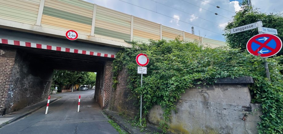 Langgasse Lahnstein Ansicht der Bahnunterführung in einer Straße, der Langgasse in Lahnstein, mit der darüber verlaufenden Bahnstrecke. Über der Unterführung weist ein Schild auf eine maximale Durchfahrtshöhe von 3,4 Metern hin. Rechts sind die Straßenschilder „Langgasse“ und „Im Nauling“ sowie ein eingeschränktes Halteverbot zu sehen. Die Durchfahrt wird durch zwei rot-weiße Poller verengt und ist von dichtem Bewuchs sowie alten Bruchsteinmauern umgeben. Im Hintergrund zeigt sich ein wolkenreicher Himmel.