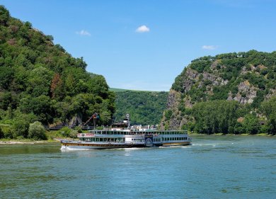 MS Goethe Das historische Schaufelradschiff „Goethe“ fährt bei sonnigem Wetter auf dem Rhein durch das UNESCO-Welterbe Oberes Mittelrheintal. Im Hintergrund erheben sich bewaldete Steilhänge und markante Felsformationen der Loreley, während sich das Schiff ruhig seinen Weg durch den Fluss bahnt.