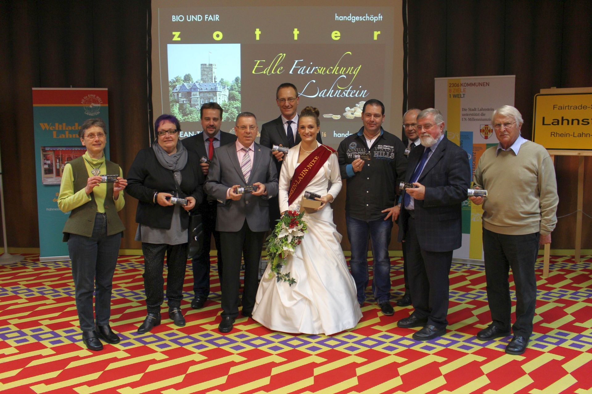 Gruppenfoto bei einer Fairtrade-Veranstaltung in Lahnstein: Mehrere Vertreterinnen und Vertreter aus Politik, Wirtschaft und Zivilgesellschaft präsentieren gemeinsam Fairtrade-Schokolade. In der Mitte steht die Lahnsteiner Weinkönigin mit Schärpe und Blumenstrauß. Im Hintergrund sind Informationsstände des Weltladens sowie eine Präsentation zur „Edlen Fairsuchung Lahnstein“ zu sehen. Rechts steht ein Fairtrade-Stadt-Schild der Stadt Lahnstein.