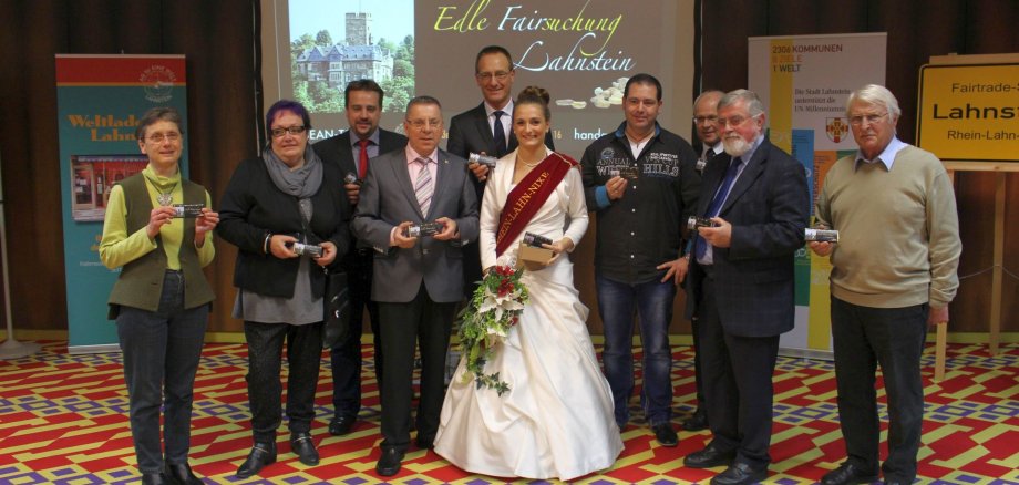 Neue faire Stadtschokolade von Lahnstein Gruppenfoto bei einer Fairtrade-Veranstaltung in Lahnstein: Mehrere Vertreterinnen und Vertreter aus Politik, Wirtschaft und Zivilgesellschaft präsentieren gemeinsam Fairtrade-Schokolade. In der Mitte steht die Lahnsteiner Weinkönigin mit Schärpe und Blumenstrauß. Im Hintergrund sind Informationsstände des Weltladens sowie eine Präsentation zur „Edlen Fairsuchung Lahnstein“ zu sehen. Rechts steht ein Fairtrade-Stadt-Schild der Stadt Lahnstein.
