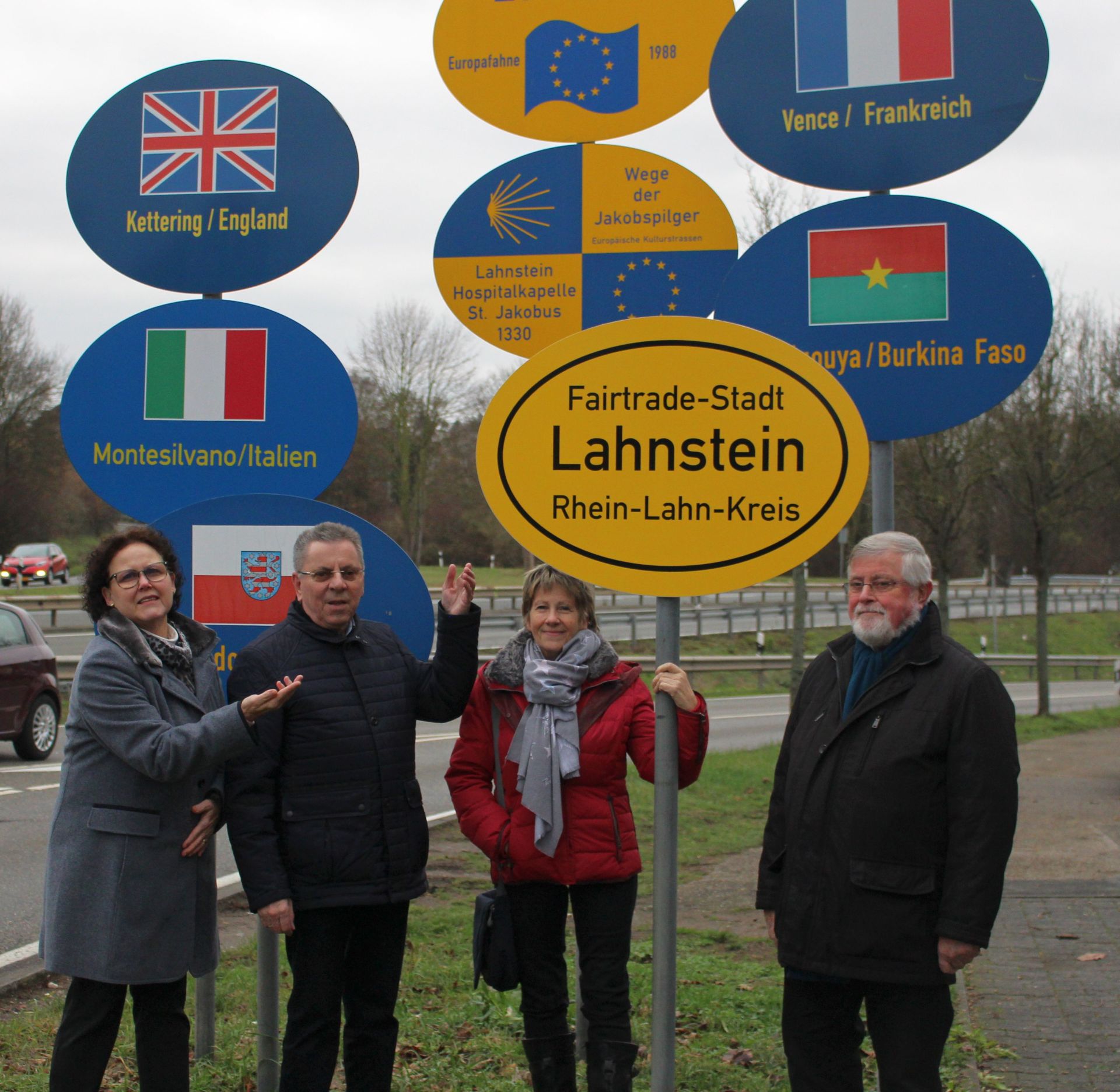 Vier Mitglieder der Fairtrade-Steuerungsgruppe stehen an einem Kreisverkehr in Lahnstein vor einem neu aufgestellten Fairtrade-Stadt-Schild. Umgeben ist das Schild von Wegweisern zu den Partnerstädten Kettering, Vence, Montesilvano und Ouagadougou sowie Hinweisen zum Jakobsweg. Die Beteiligten präsentieren die neue Beschilderung, die das Engagement Lahnsteins für fairen Handel sichtbar macht.