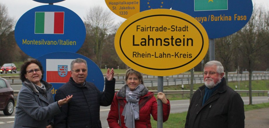 Lahnstein wird als Fairtrade-Stadt sichtbar Vier Mitglieder der Fairtrade-Steuerungsgruppe stehen an einem Kreisverkehr in Lahnstein vor einem neu aufgestellten Fairtrade-Stadt-Schild. Umgeben ist das Schild von Wegweisern zu den Partnerstädten Kettering, Vence, Montesilvano und Ouagadougou sowie Hinweisen zum Jakobsweg. Die Beteiligten präsentieren die neue Beschilderung, die das Engagement Lahnsteins für fairen Handel sichtbar macht.