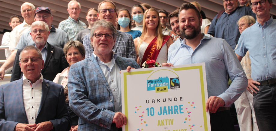 10 Jahre Fairtrade-Stadt Lahnstein Gruppenfoto mit zahlreichen Mitgliedern der Lahnsteiner Fairtrade-Steuerungsgruppe sowie Unterstützerinnen und Unterstützern. Im Vordergrund halten zwei Männer eine Urkunde zum zehnjährigen Jubiläum der Fairtrade-Stadt Lahnstein.