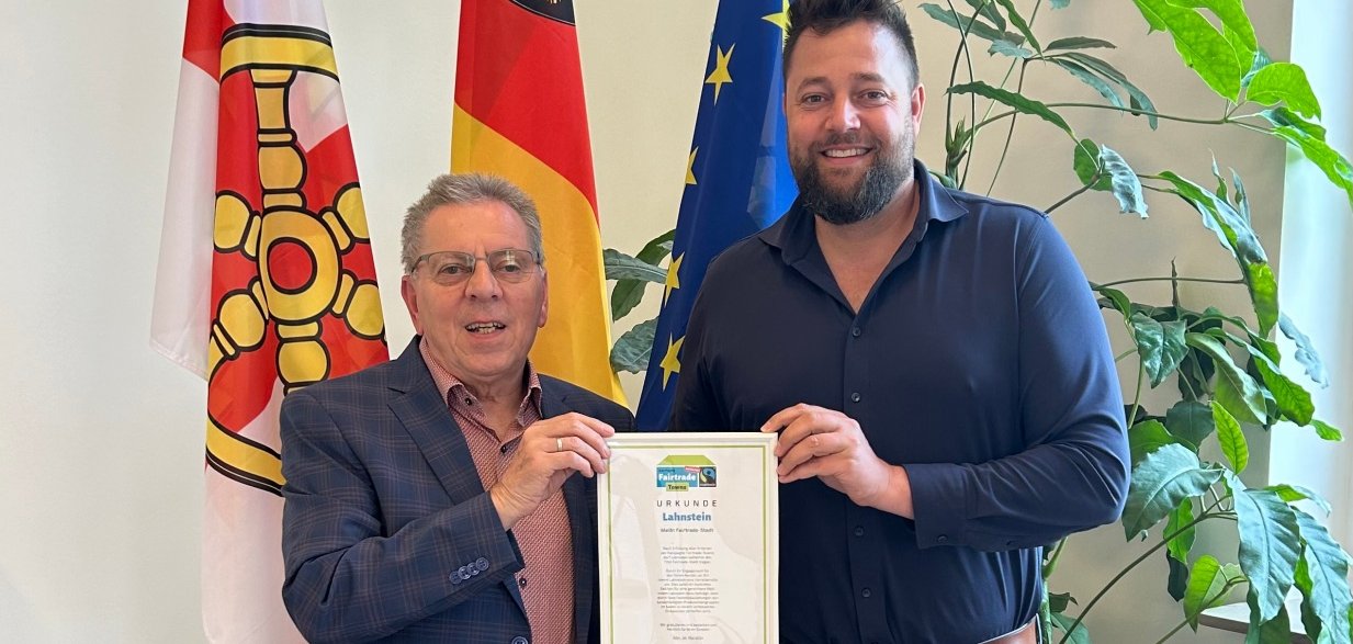 Zwei Männer stehen vor den Flaggen von Lahnstein, Rheinland-Pfalz und der Europäischen Union und halten gemeinsam eine Fairtrade-Urkunde. Neben ihnen steht eine große Zimmerpflanze.