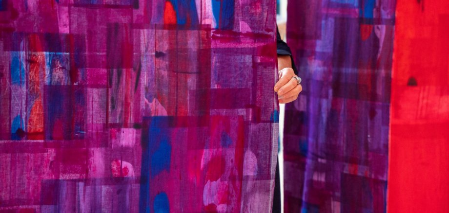 Ausstellung im Theater Lahnstein Nahaufnahme mehrerer großformatiger Stoffbahnen mit abstrakten Mustern in kräftigen Violett-, Pink-, Blau- und Rottönen. Zwischen zwei Stoffbahnen ist eine Hand zu sehen, die den Stoff zur Seite hält.