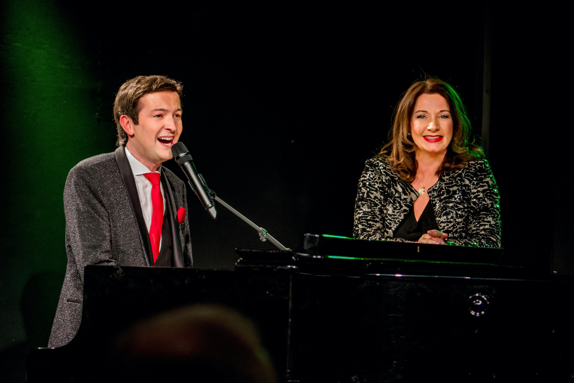 Alex Parker und Gabriela Benesch stehen vor schwarzem Hintergrund an einem Flügel.