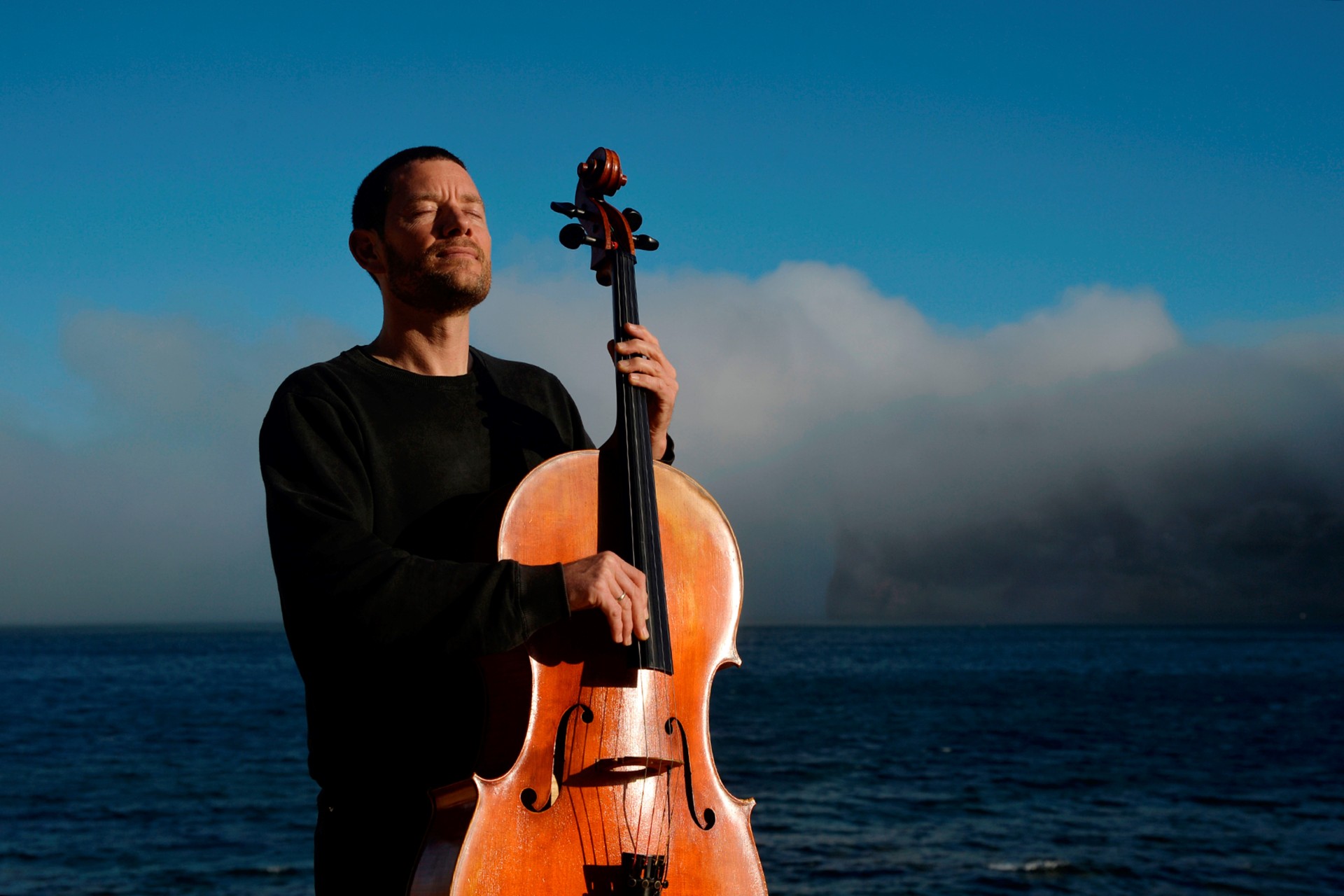Ein Mann in schwarzer Kleidung steht mit einem Cello am Meer. Er hält das Instrument aufrecht vor sich und hat die Augen geschlossen. Im Hintergrund sind das Wasser, ein blauer Himmel und eine große Wolkenformation zu sehen.