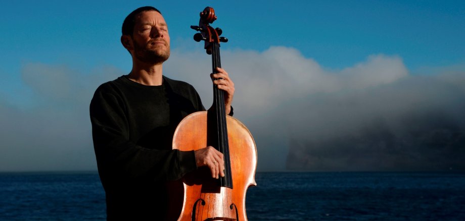 Matthieu Saglio am Cello Ein Mann in schwarzer Kleidung steht mit einem Cello am Meer. Er hält das Instrument aufrecht vor sich und hat die Augen geschlossen. Im Hintergrund sind das Wasser, ein blauer Himmel und eine große Wolkenformation zu sehen.