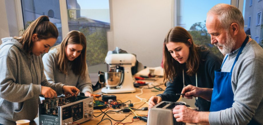 Repair Café im Jugendkulturzentrum In einem hellen Raum arbeiten vier Menschen unterschiedlicher Generationen an einem langen Holztisch zusammen. Im Vordergrund repariert ein älterer Mann in einer blauen Schürze einen Toaster, während junge Frauen daneben ein elektronisches Gerät untersuchen. Auf den Tischen liegen Werkzeuge, Kabel und Messgeräte.