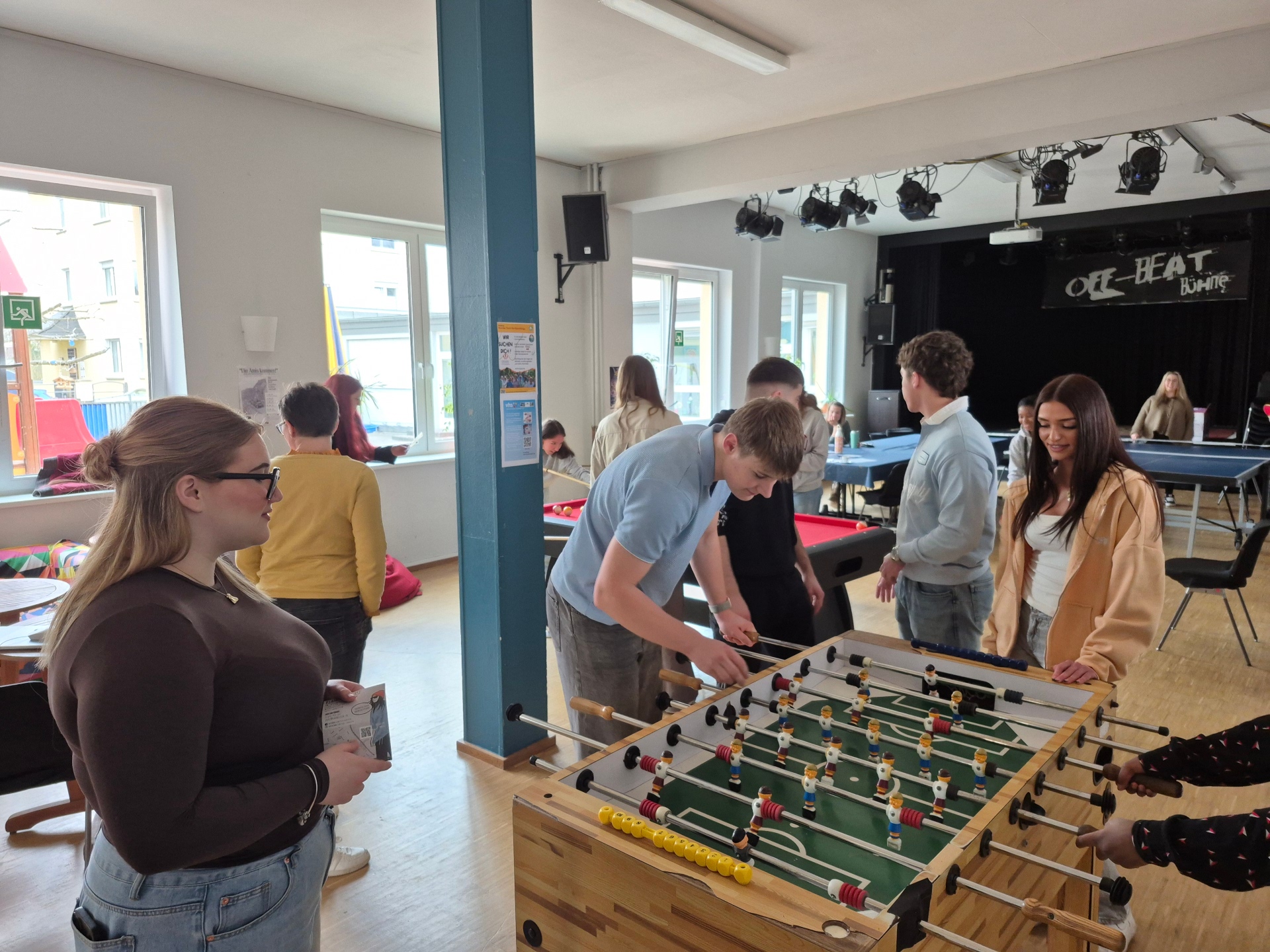 Mehrere Jugendliche stehen im Jugendkulturzentrum Lahnstein um einen Kickertisch herum und spielen gemeinsam. Im Hintergrund sind weitere junge Menschen an einem Billardtisch sowie eine kleine Bühne zu sehen.