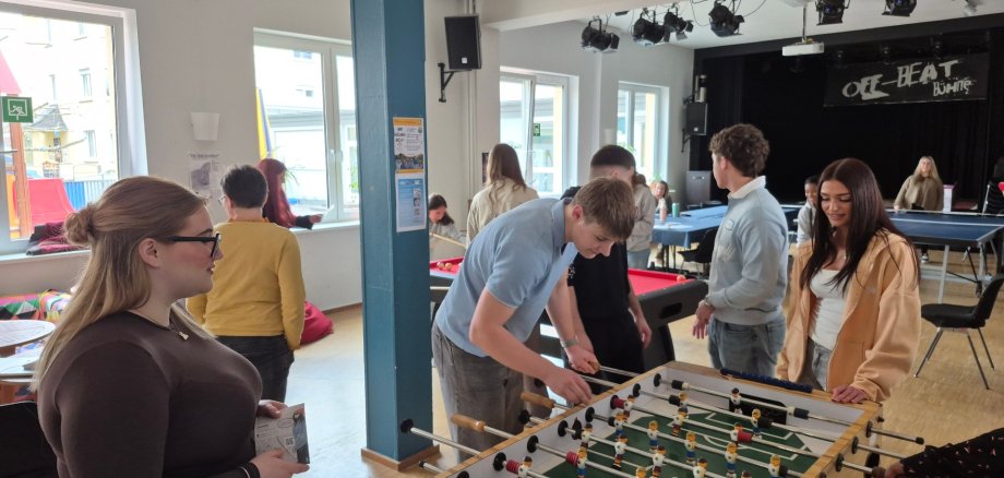 Kooperation zwischen BBS Lahnstein und Jugendkulturzentrum stärkt Praxisnähe Mehrere Jugendliche stehen im Jugendkulturzentrum Lahnstein um einen Kickertisch herum und spielen gemeinsam. Im Hintergrund sind weitere junge Menschen an einem Billardtisch sowie eine kleine Bühne zu sehen.