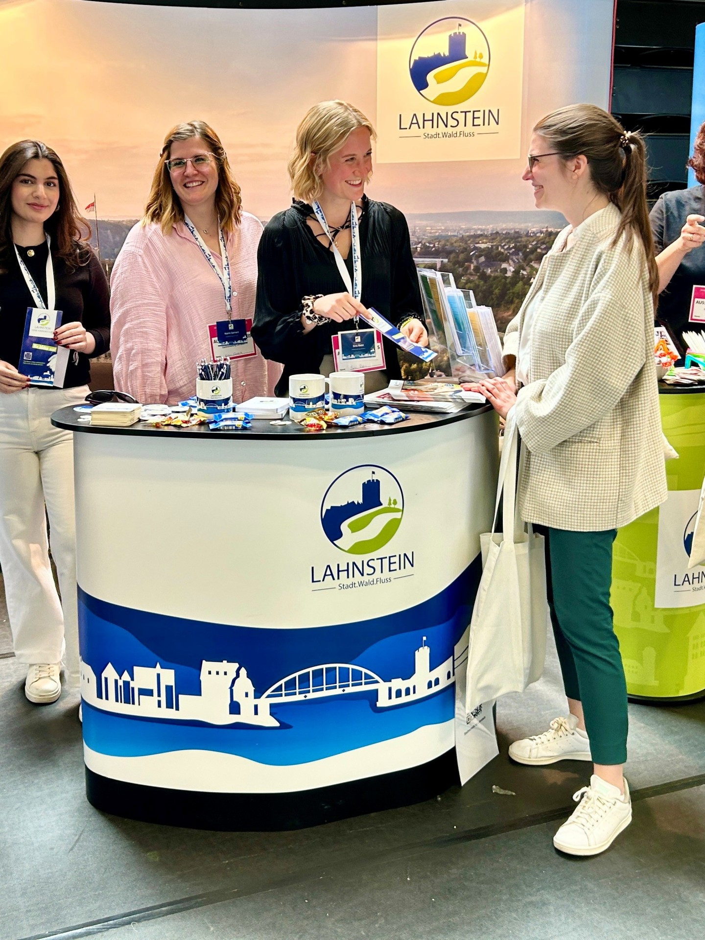 Drei Mitarbeiterinnen der Stadtverwaltung Lahnstein stehen an einem Messestand und beraten eine Besucherin. Sie halten Informationsmaterial bereit und lächeln im Gespräch. Der Stand ist mit dem Lahnstein-Logo sowie Broschüren, Kugelschreibern und kleinen Giveaways ausgestattet. Im Hintergrund ist ein großes Banner mit Stadtmotiv und der Aufschrift „Lahnstein – Stadt.Wald.Fluss“ zu sehen.