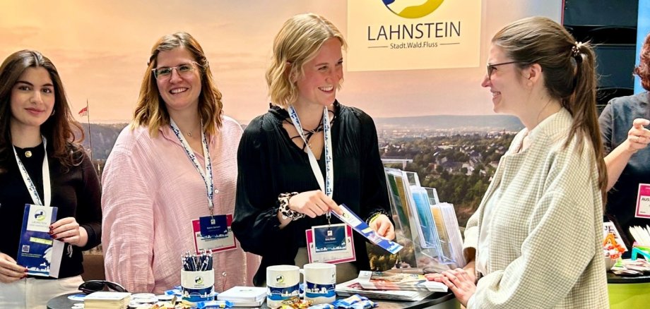 Drei Mitarbeiterinnen der Stadtverwaltung Lahnstein stehen an einem Messestand und beraten eine Besucherin. Sie halten Informationsmaterial bereit und lächeln im Gespräch. Der Stand ist mit dem Lahnstein-Logo sowie Broschüren, Kugelschreibern und kleinen Giveaways ausgestattet. Im Hintergrund ist ein großes Banner mit Stadtmotiv und der Aufschrift „Lahnstein – Stadt.Wald.Fluss“ zu sehen.