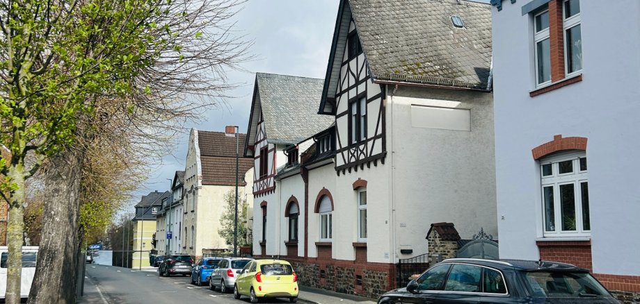 Ruhige Wohnstraße mit parkenden Autos entlang des Fahrbahnrandes. Rechts stehen mehrere Häuser mit teils Fachwerkfassaden, links säumen Bäume den Gehweg, von denen einer frisch austreibt. Über der Straße spannt sich ein blauer Himmel mit einigen Wolken.