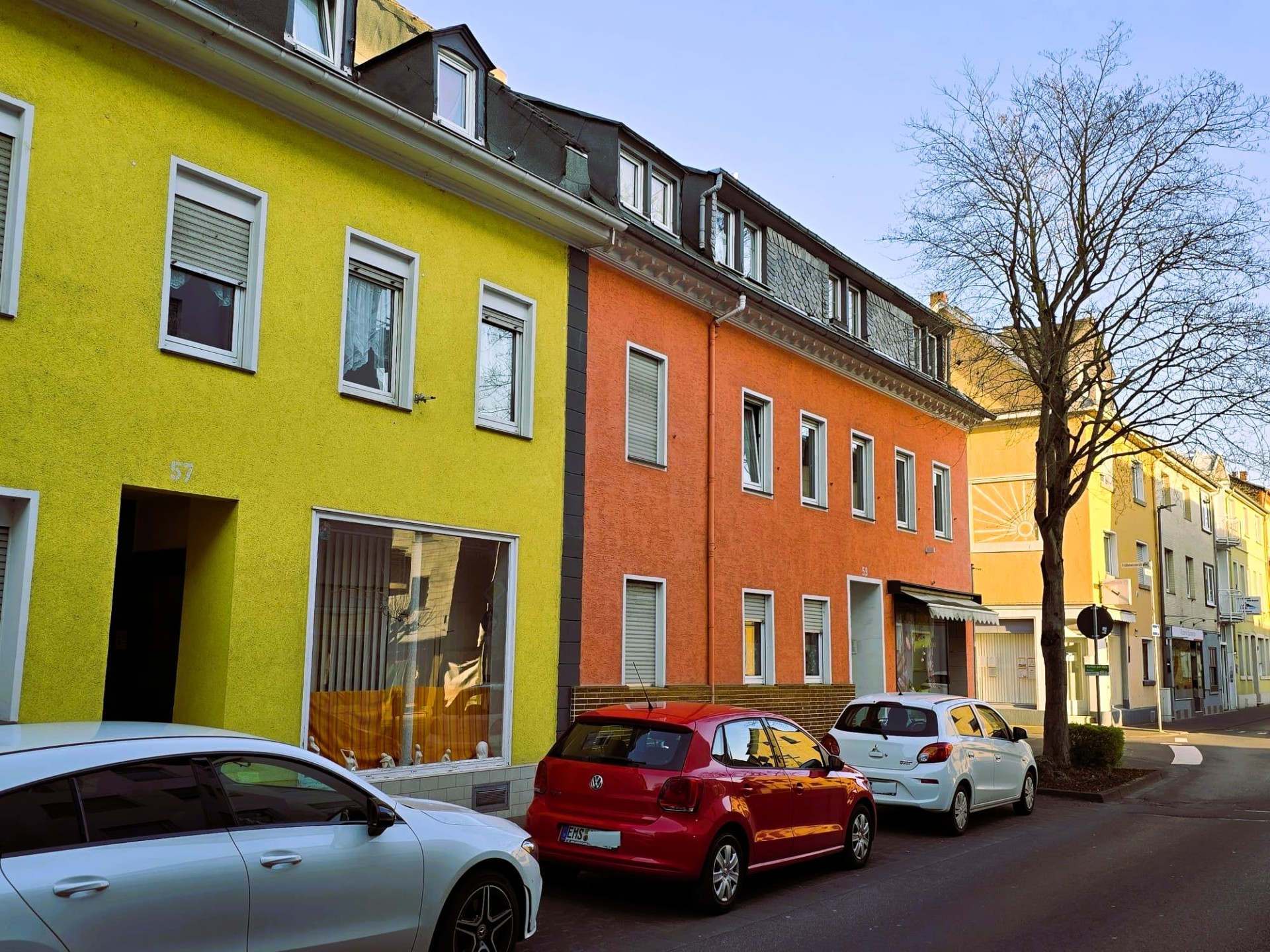 Farbfoto einer Straße mit bunt gestrichenen Häusern in Gelb und Orange, parkenden Autos am Straßenrand und einem kahlen Baum im Vordergrund.
