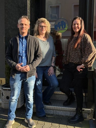 Drei Personen stehen lächelnd vor dem Eingang eines Geschäftes in Lahnstein. Links ein Mann in Jeans und dunkler Strickjacke, in der Mitte eine Frau mit Brille und gemustertem Oberteil und rechts eine Frau in einer Bluse mit Leopardenmuster. Im Hintergrund ist an der Glasscheibe ein runder, blau-grüner Aufkleber mit der Aufschrift „40 Jahre in Lahnstein“ zu sehen.