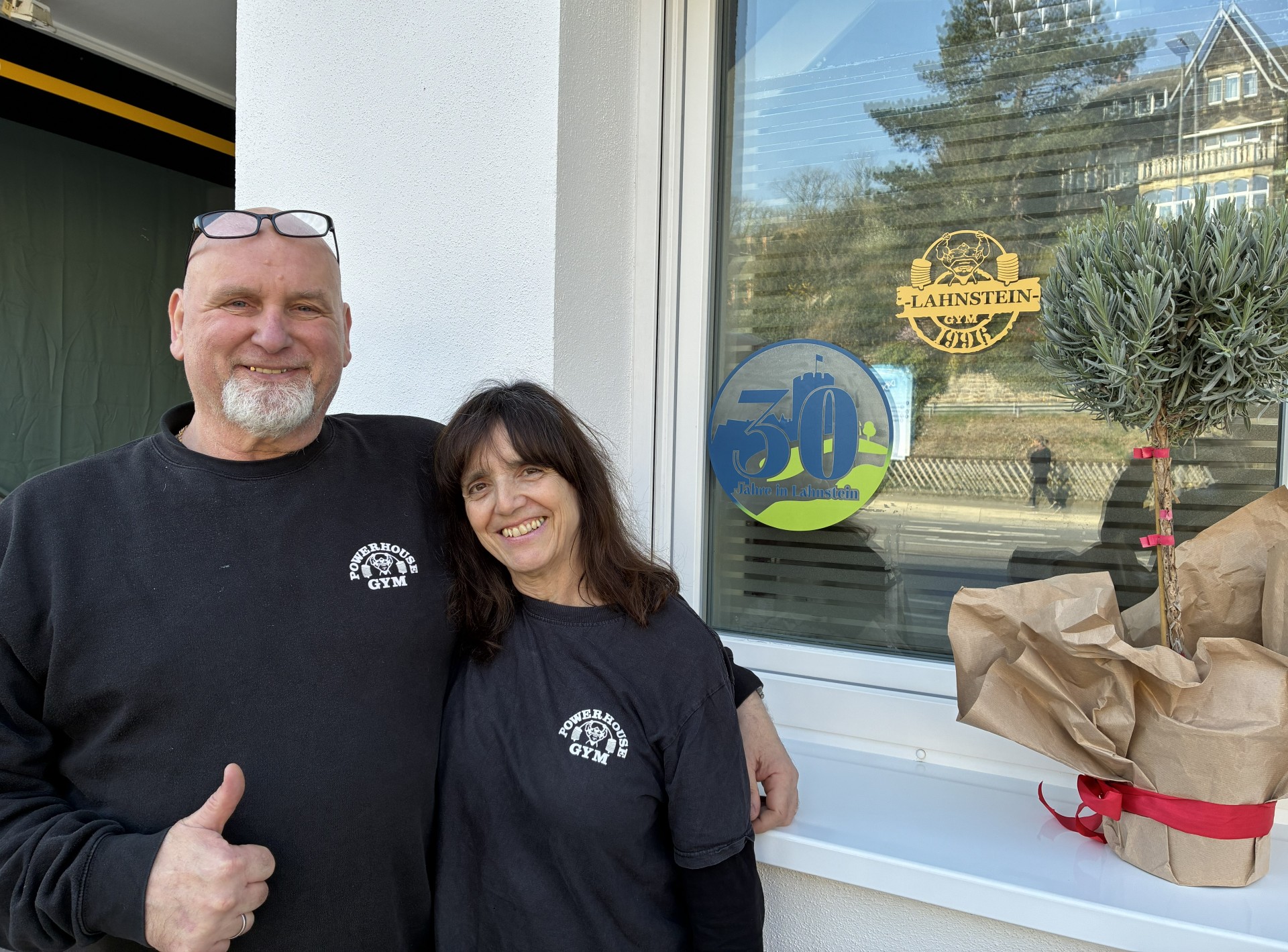 Ein lächelndes Paar steht Arm in Arm vor dem Fenster des „Powerhouse Gym“. Beide tragen schwarze T-Shirts mit dem Logo des Fitnessstudios. Der Mann zeigt einen Daumen hoch. Am Fenster klebt ein runder Jubiläumsaufkleber mit der Zahl „30“ und dem Text „Jahre in Lahnstein“. Rechts im Bild steht eine eingepackte Pflanze auf der Fensterbank.