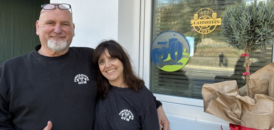 Ein lächelndes Paar steht Arm in Arm vor dem Fenster des „Powerhouse Gym“. Beide tragen schwarze T-Shirts mit dem Logo des Fitnessstudios. Der Mann zeigt einen Daumen hoch. Am Fenster klebt ein runder Jubiläumsaufkleber mit der Zahl „30“ und dem Text „Jahre in Lahnstein“. Rechts im Bild steht eine eingepackte Pflanze auf der Fensterbank.