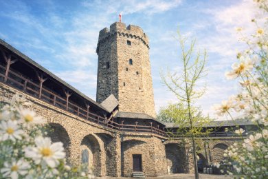"Grüner Balkon" in Lahnstein Stilisiertes Bild eines mittelalterlichen Wehrturms mit Wehrgang. Im Vordergrund sind Blumen mit weißen Blüten zu sehen, rechts im Bild ist ein Baum mit dünnen Ästen und hellgrünen Blättern.
