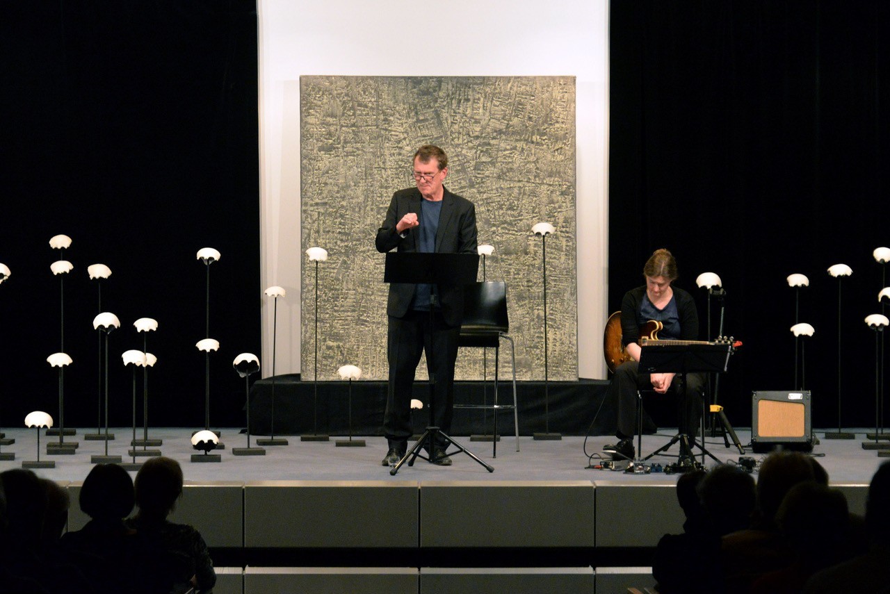 Ein Mann steht auf einer Bühne, rechts von ihm sitzt eine Frau mit einer Gitarre in der Hand