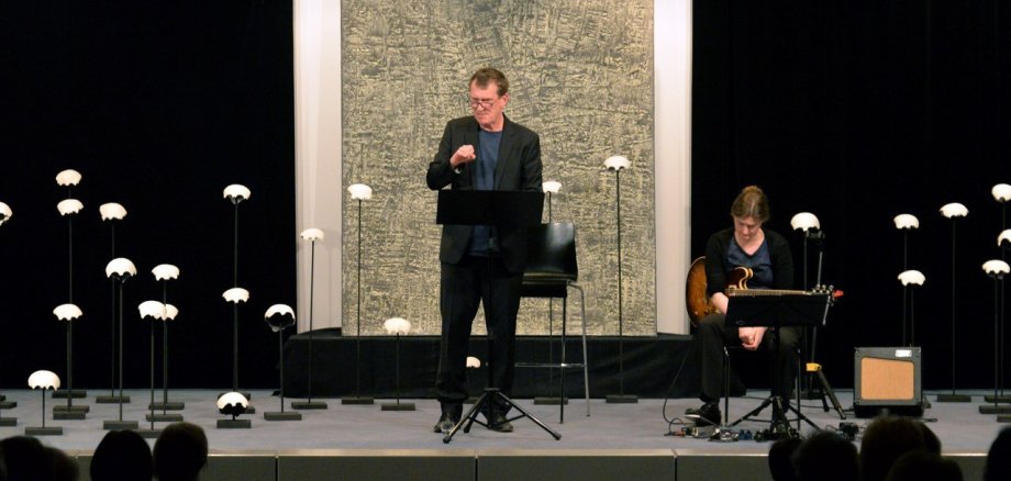 Ein Mann steht auf einer Bühne, rechts von ihm sitzt eine Frau mit einer Gitarre in der Hand