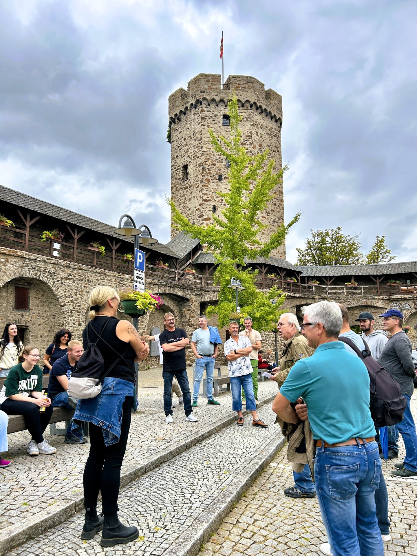 Eine Gruppe von Menschen steht und sitzt auf einem gepflasterten Platz. Im Hintergrund erhebt sich ein runder, mittelalterlicher Turm mit Fahne, umgeben von einer alten Stadtmauer mit Holzbalkonen und Blumenschmuck. Der Himmel ist bewölkt.