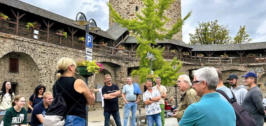 Eine Gruppe von Menschen steht und sitzt auf einem gepflasterten Platz. Im Hintergrund erhebt sich ein runder, mittelalterlicher Turm mit Fahne, umgeben von einer alten Stadtmauer mit Holzbalkonen und Blumenschmuck. Der Himmel ist bewölkt.