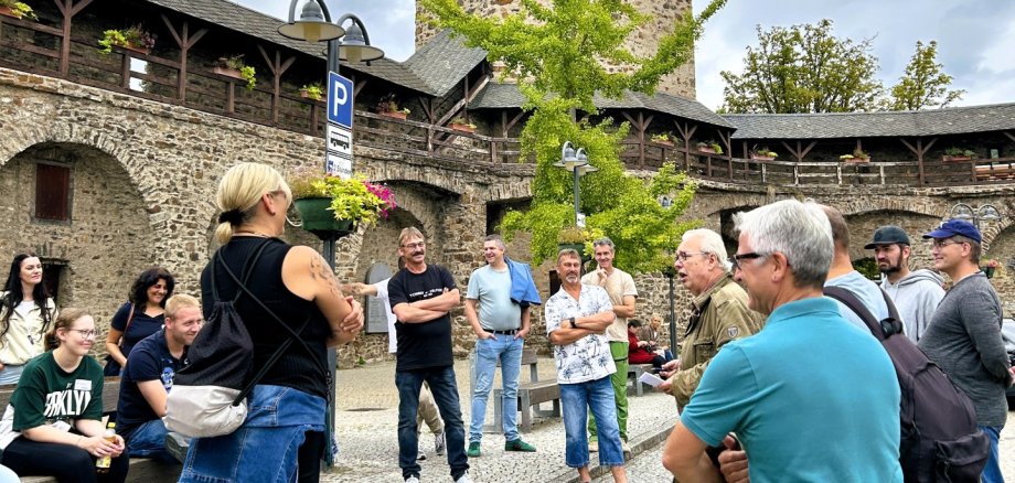 Eine Gruppe von Menschen steht und sitzt auf einem gepflasterten Platz. Im Hintergrund erhebt sich ein runder, mittelalterlicher Turm mit Fahne, umgeben von einer alten Stadtmauer mit Holzbalkonen und Blumenschmuck. Der Himmel ist bewölkt.