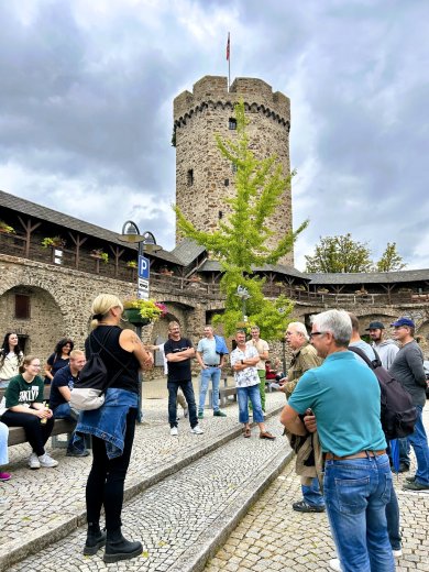 Eine Gruppe von Menschen steht und sitzt auf einem gepflasterten Platz. Im Hintergrund erhebt sich ein runder, mittelalterlicher Turm mit Fahne, umgeben von einer alten Stadtmauer mit Holzbalkonen und Blumenschmuck. Der Himmel ist bewölkt.