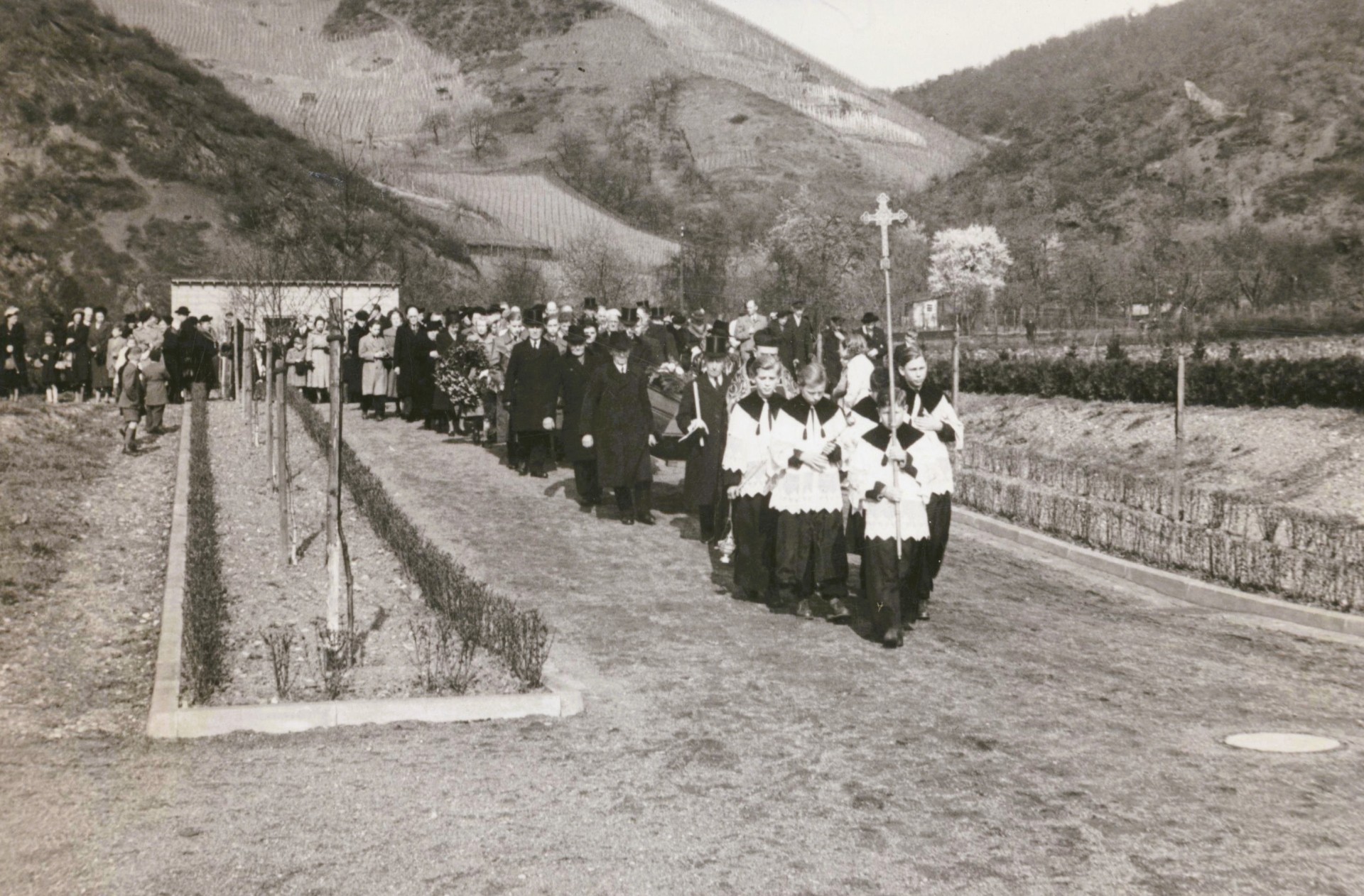 Schwarzweißfoto einer Trauerprozession auf einem Friedhof: Ministranten mit Kreuz gehen voran, dahinter folgen Geistliche und zahlreiche Trauergäste, während ein Sarg getragen wird; im Hintergrund sind Weinberge an Hängen zu sehen.