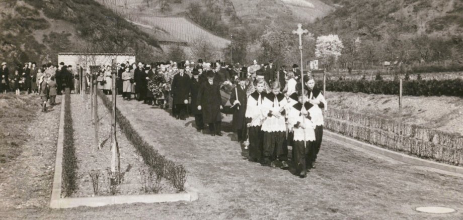 Schwarzweißfoto einer Trauerprozession auf einem Friedhof: Ministranten mit Kreuz gehen voran, dahinter folgen Geistliche und zahlreiche Trauergäste, während ein Sarg getragen wird; im Hintergrund sind Weinberge an Hängen zu sehen.