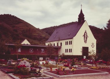 Farbfoto eines Friedhofs mit gepflegten Gräbern und Blumenbeeten vor einer kleinen Kirche mit Turm; dahinter erheben sich bewaldete Hügel unter bewölktem Himmel.