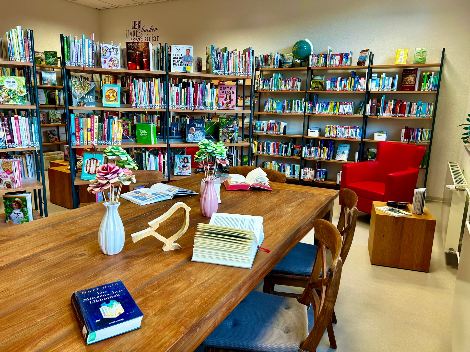 Ein gemütlicher Bibliotheksraum mit mehreren Bücherregalen voller Bücher an den Wänden. In der Mitte steht ein großer Holztisch mit offenen Büchern, kleinen Vasen mit dekorativen Blumen und einigen Stühlen. Rechts befindet sich ein roter Sessel neben einem kleinen Beistelltisch.