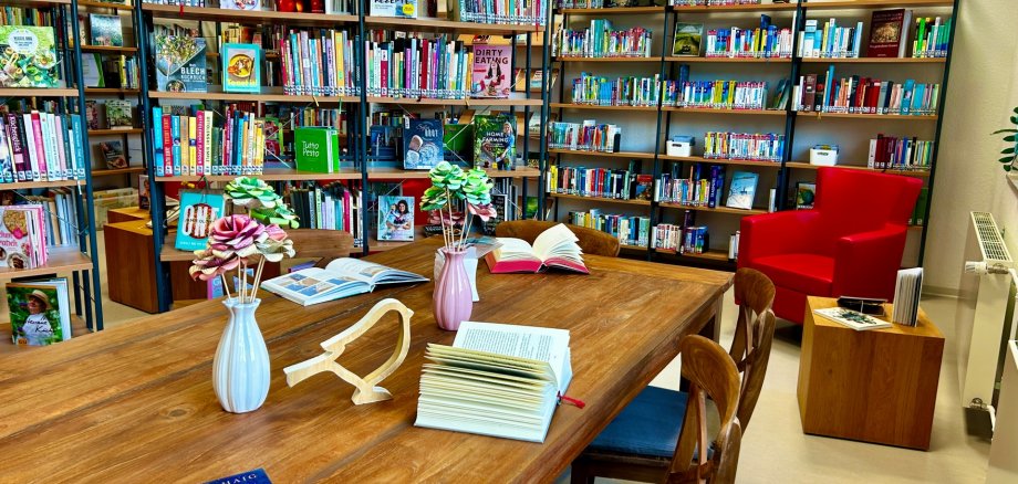 Ein gemütlicher Bibliotheksraum mit mehreren Bücherregalen voller Bücher an den Wänden. In der Mitte steht ein großer Holztisch mit offenen Büchern, kleinen Vasen mit dekorativen Blumen und einigen Stühlen. Rechts befindet sich ein roter Sessel neben einem kleinen Beistelltisch.