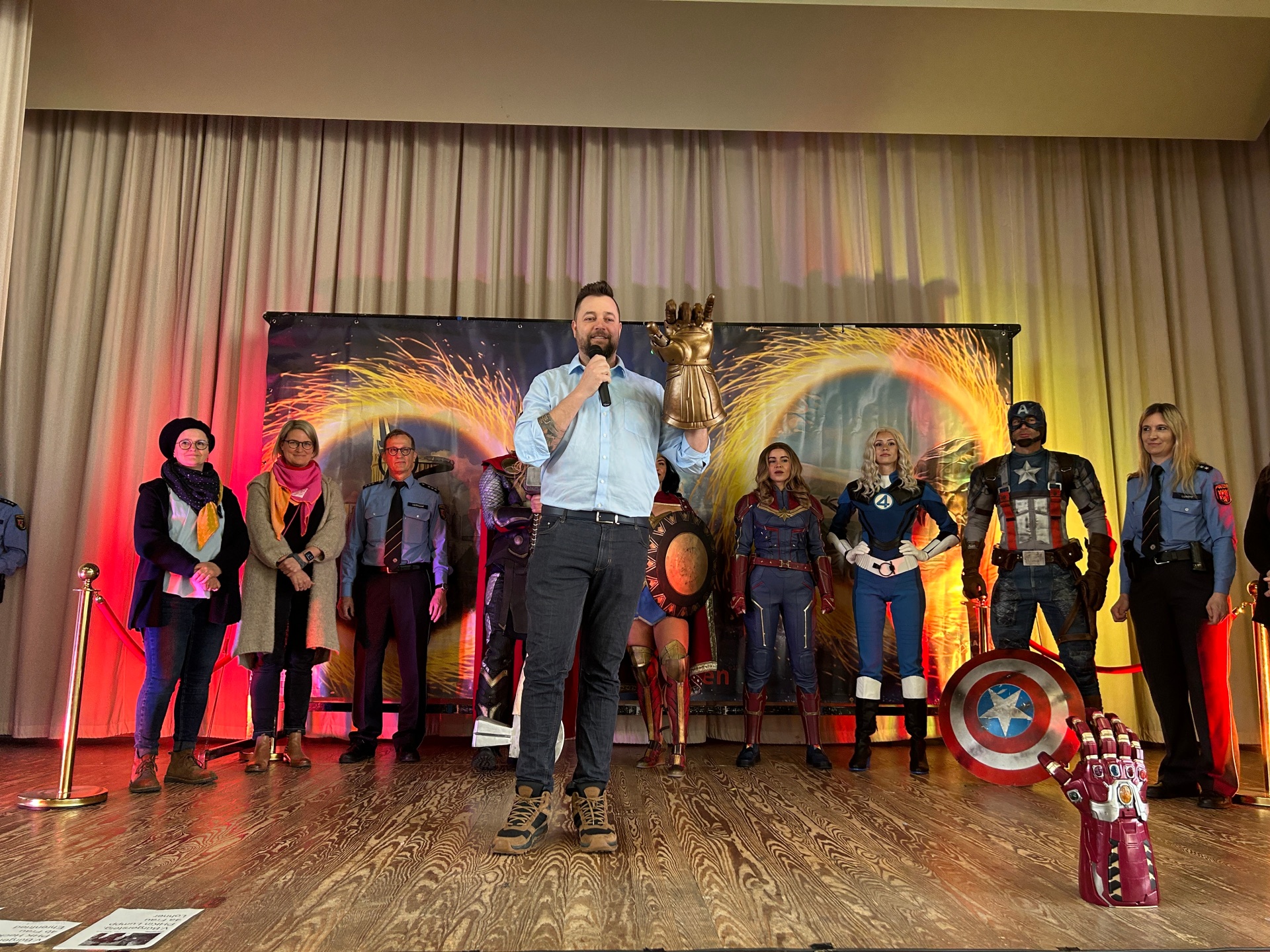 Oberbürgermeister steht auf einer Bühne und spricht ins Mikrofon, während er einen großen goldenen Handschuh hochhält. Hinter ihm stehen verkleidete Superhelden wie Captain America, Captain Marvel und weitere Figuren sowie mehrere Polizeibeamte mit und ohne Uniform. Die Bühne ist mit bunten Lichteffekten dekoriert.