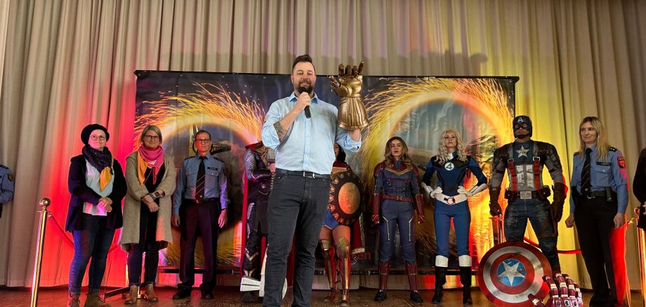 Oberbürgermeister steht auf einer Bühne und spricht ins Mikrofon, während er einen großen goldenen Handschuh hochhält. Hinter ihm stehen verkleidete Superhelden wie Captain America, Captain Marvel und weitere Figuren sowie mehrere Polizeibeamte mit und ohne Uniform. Die Bühne ist mit bunten Lichteffekten dekoriert.