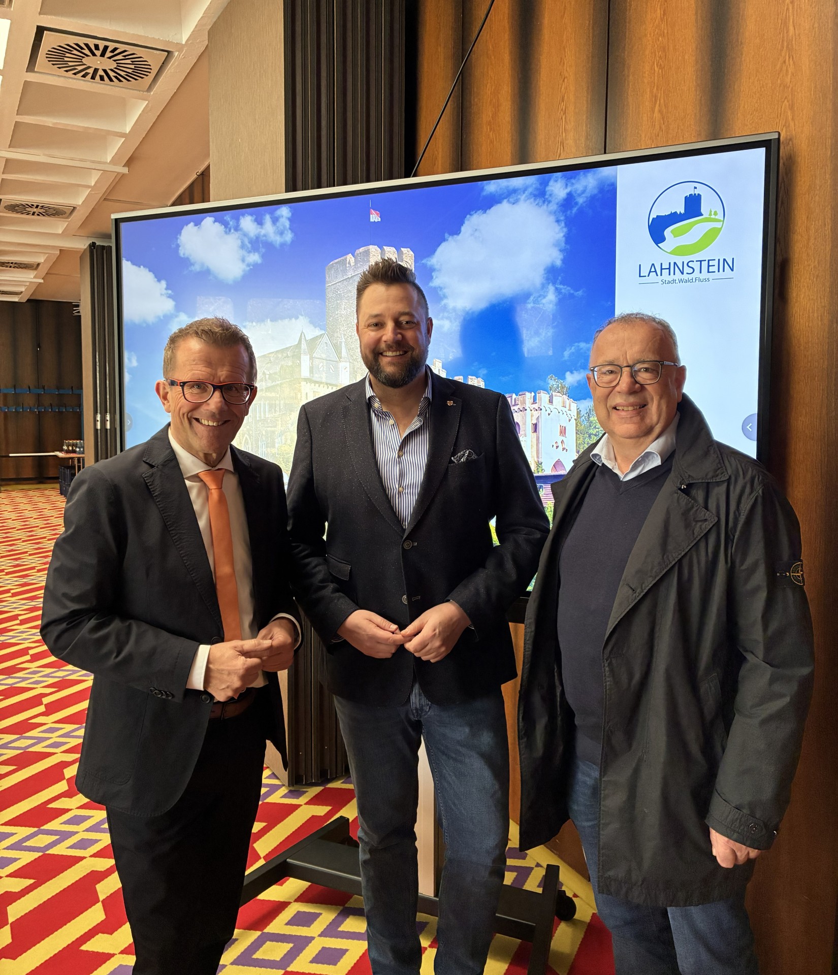 Drei lächelnde Männer in Business-Kleidung stehen in einem Tagungsraum vor einem großen Bildschirm. Auf dem Display ist eine Luftaufnahme einer Burg in Lahnstein unter strahlend blauem Himmel zu sehen, ergänzt durch das offizielle Logo der Stadt Lahnstein. Der Teppich im Raum hat ein markantes rot-gelbes geometrisches Muster.