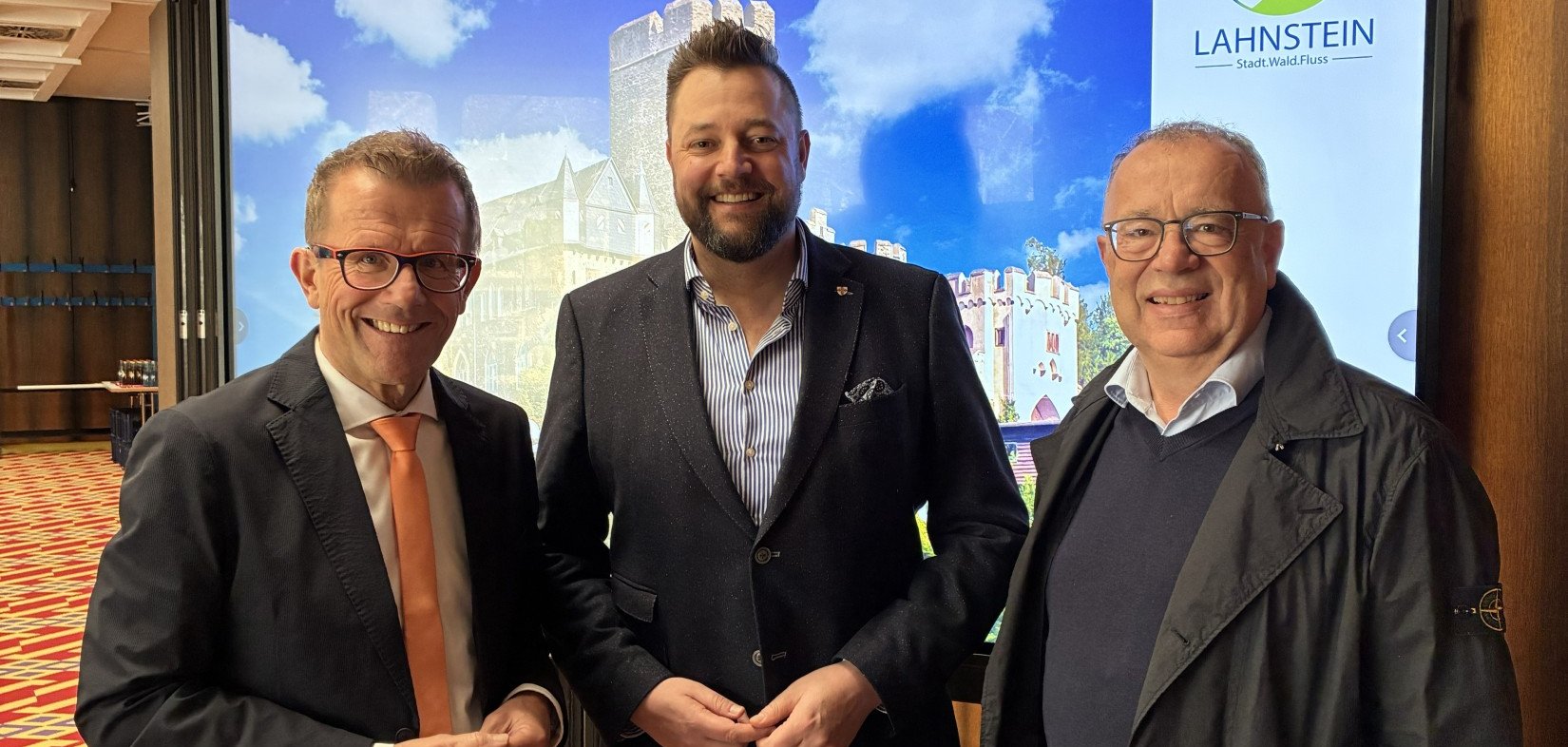 Drei lächelnde Männer in Business-Kleidung stehen in einem Tagungsraum vor einem großen Bildschirm. Auf dem Display ist eine Luftaufnahme einer Burg in Lahnstein unter strahlend blauem Himmel zu sehen, ergänzt durch das offizielle Logo der Stadt Lahnstein. Der Teppich im Raum hat ein markantes rot-gelbes geometrisches Muster.